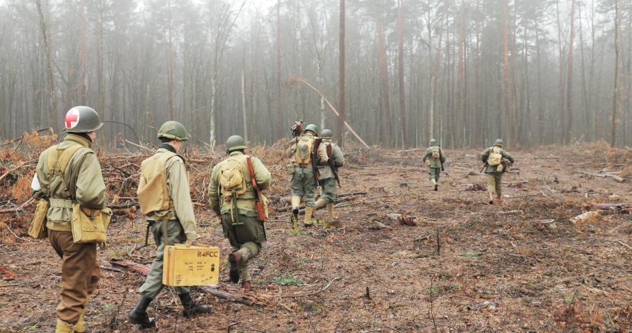 Historical Re-enactment. Re-enactors Dressed As American Soldiers Of USA Infantry Of World War II Marching Walking Along Forest Road In Autumn Day. Group of Soldiers Marching In Forest