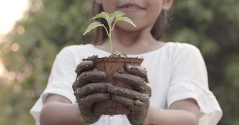 Asian child girl with mud dirty hands holding young seedlings in recycled fiber pots in the garden.Concept of child love in environmental conservation.Slow motion shot.