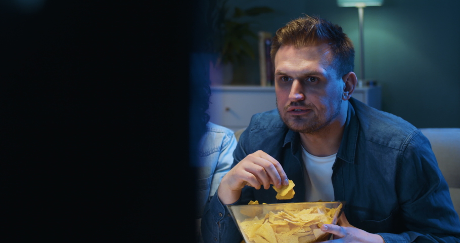 Two Caucasian young guys sitting on sofa at night in dark living room, eating potato chips and watching football match on TV. Best male friends having snacks in front of television with sport channel.