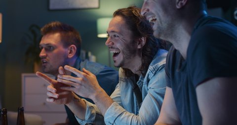 Close up of happy cheerful Caucasian men sitting on sofa in living room at night and watching comedy movie on TV. Joyful guys laughing. Male best friends spending time together.