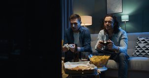 Handsome Caucasian joyful men friends sitting on sofa in tension and worrying while playing videogame with joysticks in front TV screen. Videogaming at home concept.