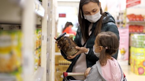 Young woman with child girl in medical masks buys a canned food in grocery department at supermarket. Virus epidemic concept.