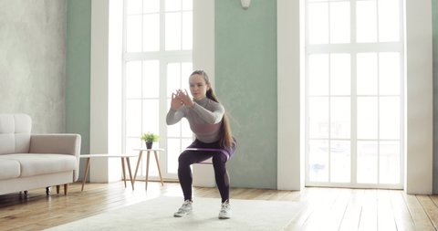 Sporty woman is training making squats and lunges at home in living room. Sporty girl having workout. Training and making physical exercises with rope, fitness elastic band. Sport concept.