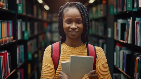 University Library: Smart Beautiful Black Girl Standing Next to Bookshelf Holding and Reading Text Book, Doing Research for Her Class Assignment and Exam Preparations. Authentic Students Study and Suc