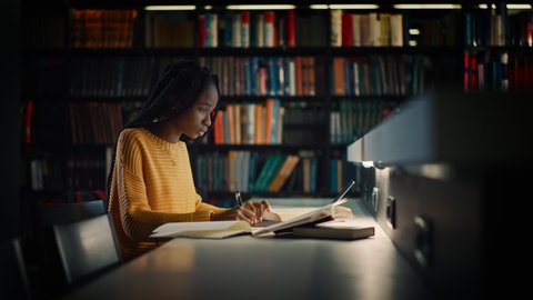 University Library: Gifted Black Girl uses Laptop, Writes Notes for the Paper, Essay, Study for Class Assignment. Students Learning, Studying for Exams College. Side View Portrait with Bookshelves