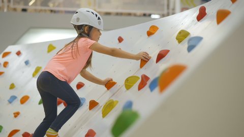 Cute kid in safety helmet climbing on small wall in attraction playground. Child training in climbing center. Girl toddler having fun in indoor amusement park