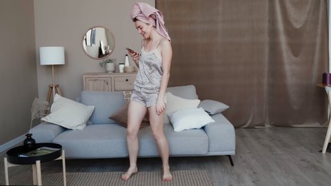 Cool girl dressed nightwear and home slippers wearing wireless headphones dancing in living room smiling looking at mobile phone hair wrapped in bath towel, morning fun background of sofa and cushions