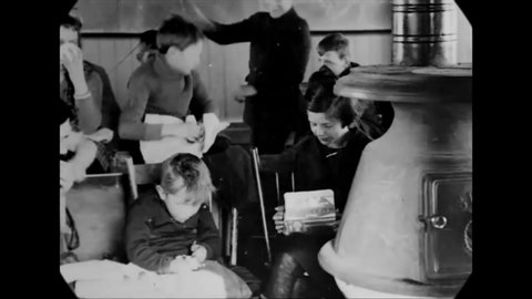 CIRCA 1923 - During lunchtime in a one-room schoolhouse in rural Ontario, Canada, children eat by the furnace while two boys have a play fight.