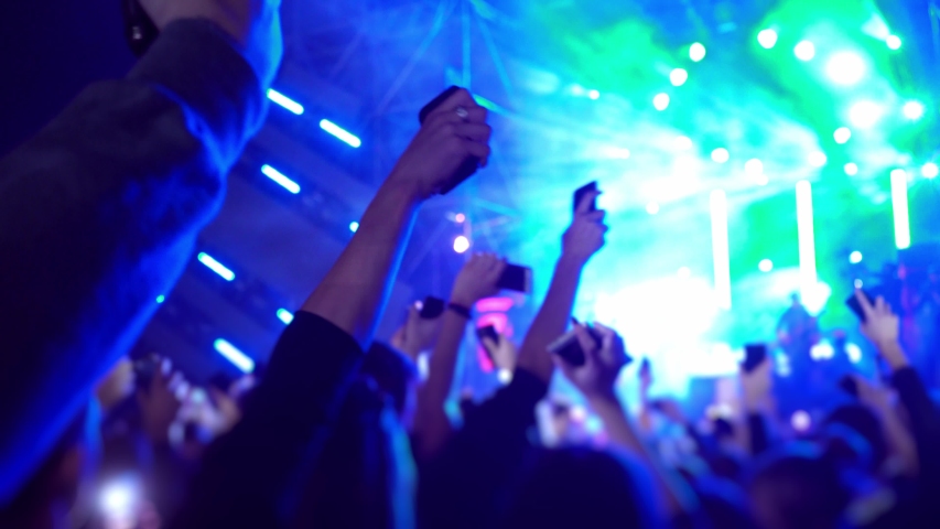 Happy people are watching an amazing musical concert. Merry fans jump and raise their hands up. Crowd of excited fans applauding to popular band performing favorite song. The crowd watches a concert. Royalty-Free Stock Footage #1049111248