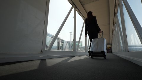 Business lady walk to airliner by jet bridge with trolley case, wide angle shot, low camera position. Boarding finishing soon, but woman go unhurried. Modern international airport, glass airgate passa