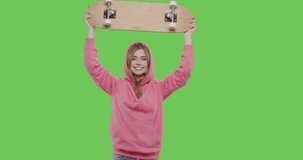 Young positive woman having fun isolated on green screen background at studio . Blonde girl dressed in pink hoodie holding skateboard. 4k raw footage video side view
