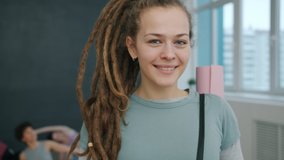 Portrait of charming young woman yoga student standing indoors in studio with mat smiling looking at camera. Training youth is visible in background. - Powered by Shutterstock - Get 15% off with code: PIKWIZARD15