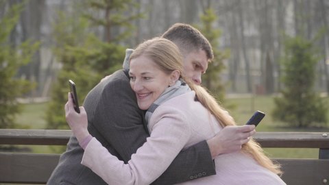 Side view of hugging Caucasian adult couple using smartphones. Blond woman and brunette man using social media on a date outdoors. Device addiction, social communication, lifestyle.