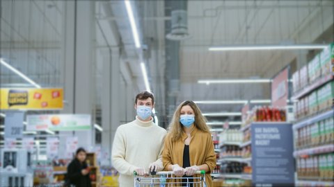 serious married couple family in medical mask with grocery cart is standing in supermarket, there crowds of customers walking around, timelapse. Protection from coronavirus. Panic, people buying up