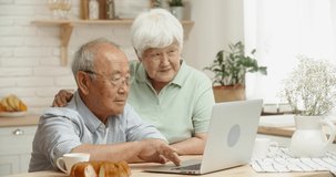 Happy asian senior couple sitting at kitchen table surfing web on laptop together, doing online shopping or watching videos and discussing - happy retirement concept 4k footage