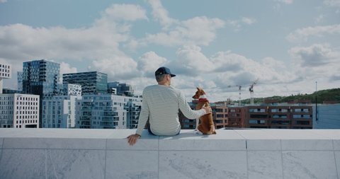 Hipster man and pet brown dog sit on edge of rooftop. Guy and best friend animal look over on empty city on sunny summer day. Social isolation concept. Peace and tranquility in middle of city buzz