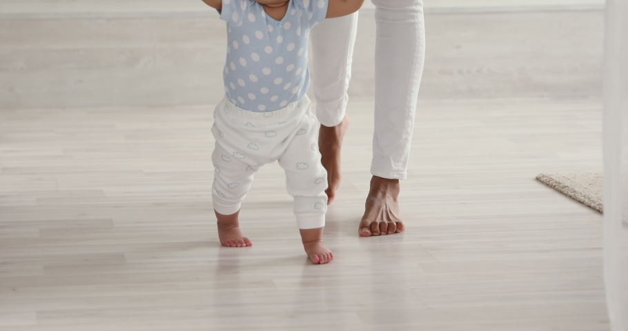 Infant barefoot baby girl boy learning to walk standing on warm floor. African american mum holding hand helping cute little child making first steps at home. Underfloor heating concept, close up view Royalty-Free Stock Footage #1049156362
