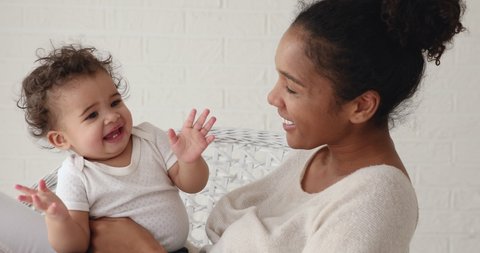 Sweet funny little mixed race infant boy son clapping hands having fun with young adult mom or nanny sitting on chair at home. Happy afro american mom holding kid playing with cute baby girl daughter.