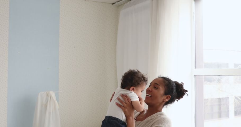 Happy young adult african american mom holding cute adorable infant child girl lifting up standing in bedroom. Cheerful adult mommy playing with cute funny kid baby boy having fun together indoors. Royalty-Free Stock Footage #1049156374