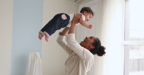 Happy young adult african american mom holding cute adorable infant child girl lifting up standing in bedroom. Cheerful adult mommy playing with cute funny kid baby boy having fun together indoors.