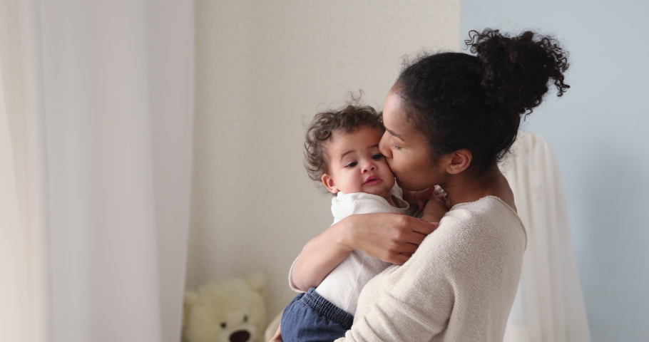 Loving tender african american young mum holding adorable cute baby girl daughter embracing kissing small kid. Happy affectionate mixed race mother cuddling with infant child boy son standing at home. | Shutterstock HD Video #1049156401
