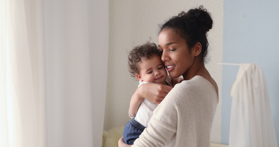 Loving tender african american young mum holding adorable cute baby girl daughter embracing kissing small kid. Happy affectionate mixed race mother cuddling with infant child boy son standing at home. Royalty-Free Stock Footage #1049156401