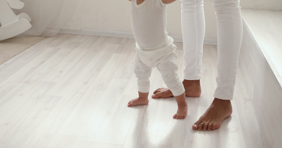 Cute barefoot small mixed race baby girl learning to walk holding mom hands. Funny adorable african infant toddler child daughter playing with mum making first step on warm heated floor. Close up view Royalty-Free Stock Footage #1049156416