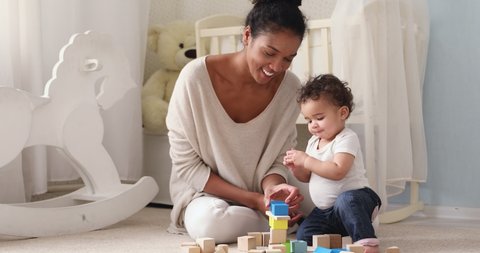 Happy african mum helping adorable baby girl daughter having fun together sitting on warm floor. Mixed race family mama or nanny teaching little infant boy son learning playing wooden blocks at home.