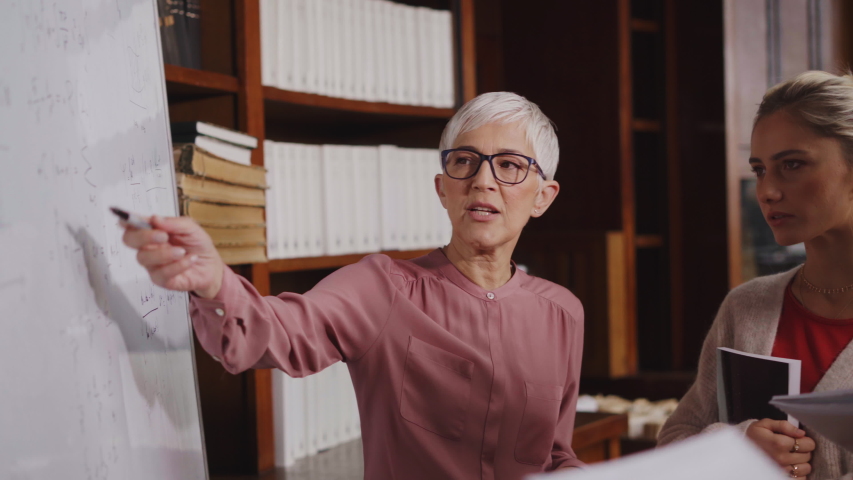 Professor helping student in library at college. Group of multiethnic students taking an active part in a lesson while standing in library. Mature woman teacher and university student discussing. - Powered by Shutterstock - Get 15% off with code: PIKWIZARD15