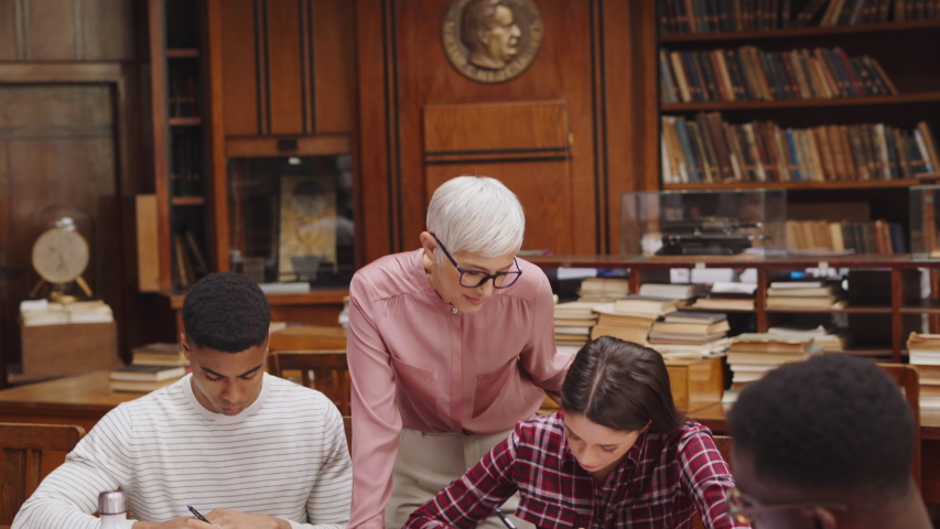 Senior woman teacher working with college students in library. Graduates consulting with their lecturer informally during a break in library. Group of students studying with their professor. Royalty-Free Stock Footage #1049156923