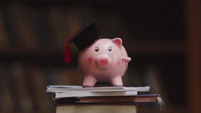Pink piggy bank wearing graduation cap and holding pencil on pile of books in college library. Student loan and debt concept. Close up of piggybank with black graduate hat. - Powered by Shutterstock - Get 15% off with code: PIKWIZARD15