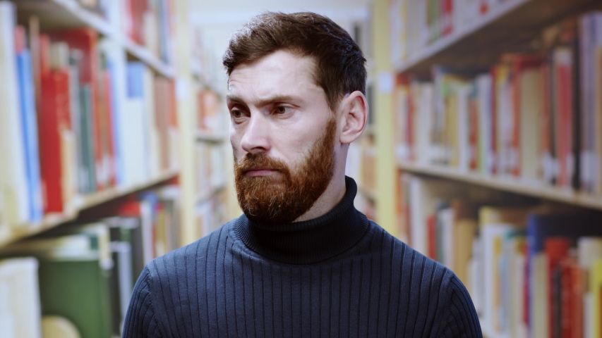 Bearded confused young man standing near bookshelves searching books shrugging shoulders cant help dont understand look at camera in the library. Royalty-Free Stock Footage #1049159647