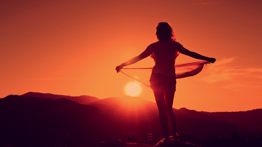 Young woman with hands up holding scarf in wind on the mountain landscape at sunset