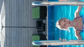beautiful blonde comes out of the pool during summer resort, thin young girl in a blue bathing suit climbing the ladder, smartphone vertical screen
