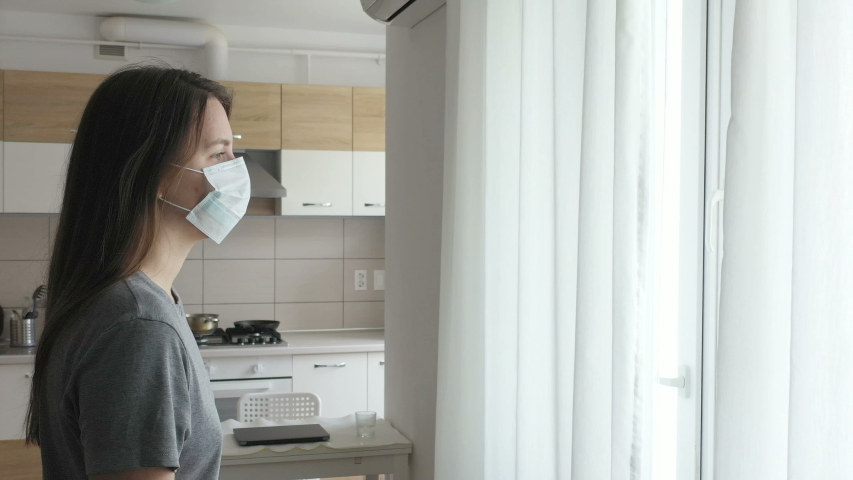 Woman in Face Mask Looking out the Window. Staying Home in Quarantine. Royalty-Free Stock Footage #1049189035