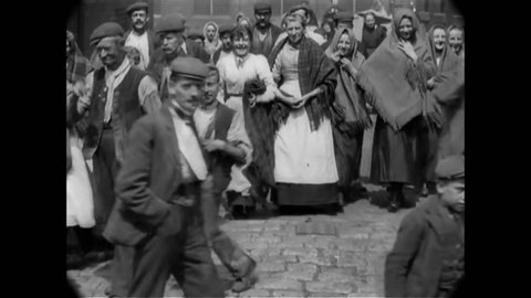 CIRCA 1901 - Men, women and children smile at the camera as they walk past on a humble road in Glebe Mills, Hollinwood, England.