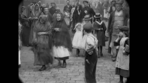 CIRCA 1900 - As adults walk past the camera, English children stop to stare and make faces.