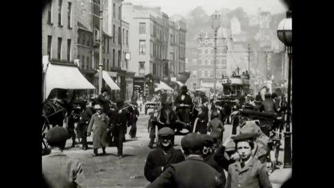 CIRCA 1902 - Pedestrians of all ages look at a camera in Ireland as horses pull carriages and trolleys through a city street.