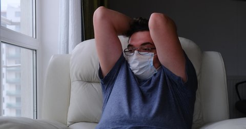 Man with medical mask sits in armchair next to a window, looks outside, runs his fingers through his hair in despair, while quarantined at home.