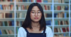 Portret of beautiful asian teen girl student with black hair and glasses looking at camera and smiling in library next to book rack.