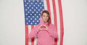 Pretty stylish girl in pink hoodie celebrating independence day and having fun over national usa flag on white wall on background. Young woman show heart sign symbol . 4k video footage slow motion