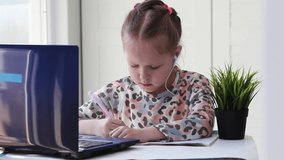 A little girl writes in a notebook and looks at a laptop.Distance learning, school online.