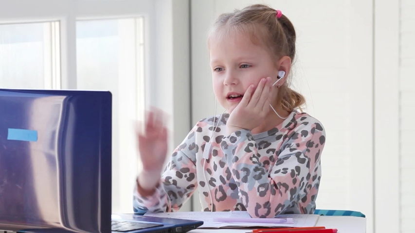 The girl in the headphones looks with interest at the laptop screen and waves hello. Distance learning, school online. Royalty-Free Stock Footage #1049212036