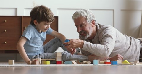Happy middle aged man lying on floor carpet, playing colorful wooden cubes with small school aged grandson in living room. Smiling different generations family enjoying weekend leisure time at home.