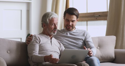 Handsome young man cuddling happy elderly mature father, helping teaching using computer applications, sitting together in living room. Middle aged dad discussing media news with grown up son indoors.