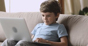 Smart little child boy sitting on sofa, holding on lap computer, using early development educational programs, studying alone at home. Addicted to technology happy small kid playing game on laptop.