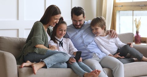 Little schoolgirl relaxing on sofa with smiling parents and younger brother, showing funny video on smartphone. Full length happy family using mobile applications, enjoying leisure time together.