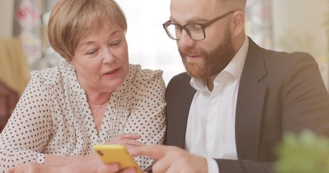 Handsome businessman teaching his mature grandmother using modern smartphone. Good looking old woman and young man looking at mobile phone screen while sitting at home