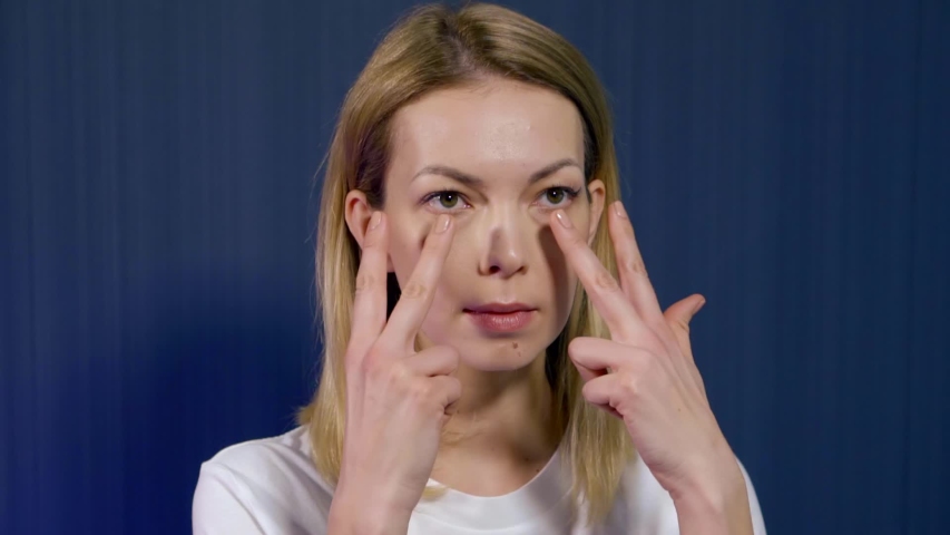 Close-up portrait of a young blonde girl who is in the Studio on a blue background, she is engaged in facial massage and facial gymnastics. Creates an area around the eyes.