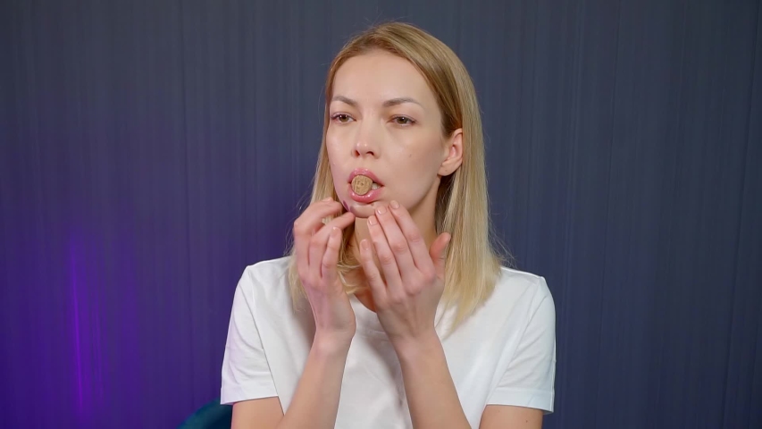 Close-up portrait of a blonde girl who is in the Studio on a blue background, she has a cork in her mouth and she is engaged in facial massage and facial gymnastics